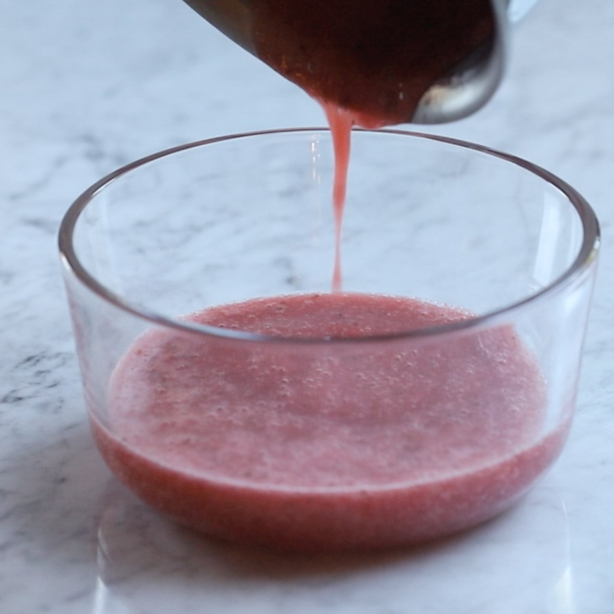 Fresh strawberry smoothie in a glass bowl, being poured from a saucepan, on a marble surface.